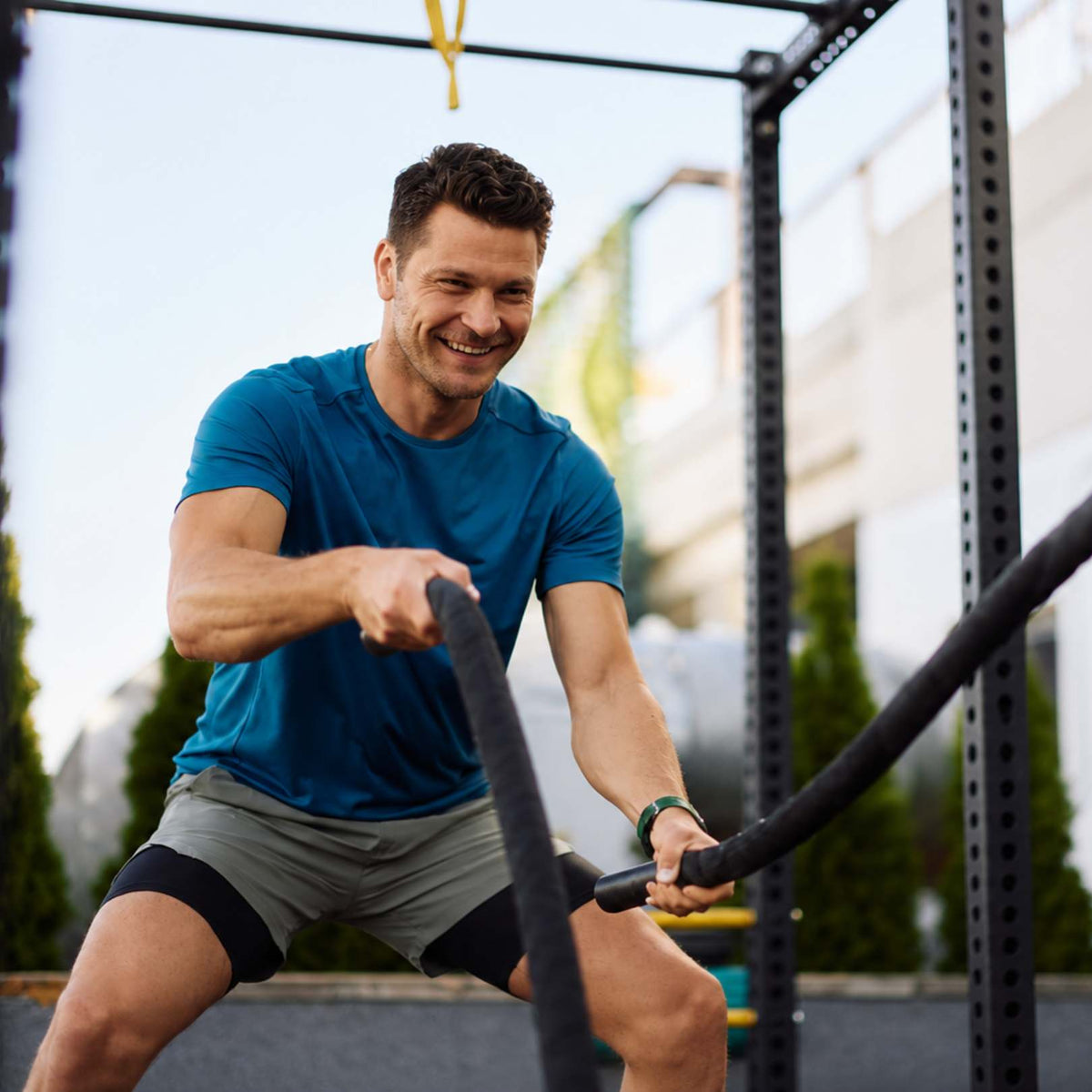 A smiling man in a blue athletic t-shirt and gray shorts exercises with battle ropes on an outdoor gym structure.