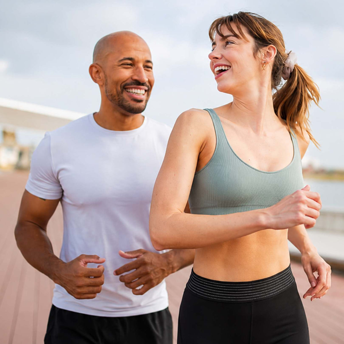 A smiling man in a white t-shirt and a woman in a sage green sports bra and black leggings jogging together outdoors on a wooden boardwalk.